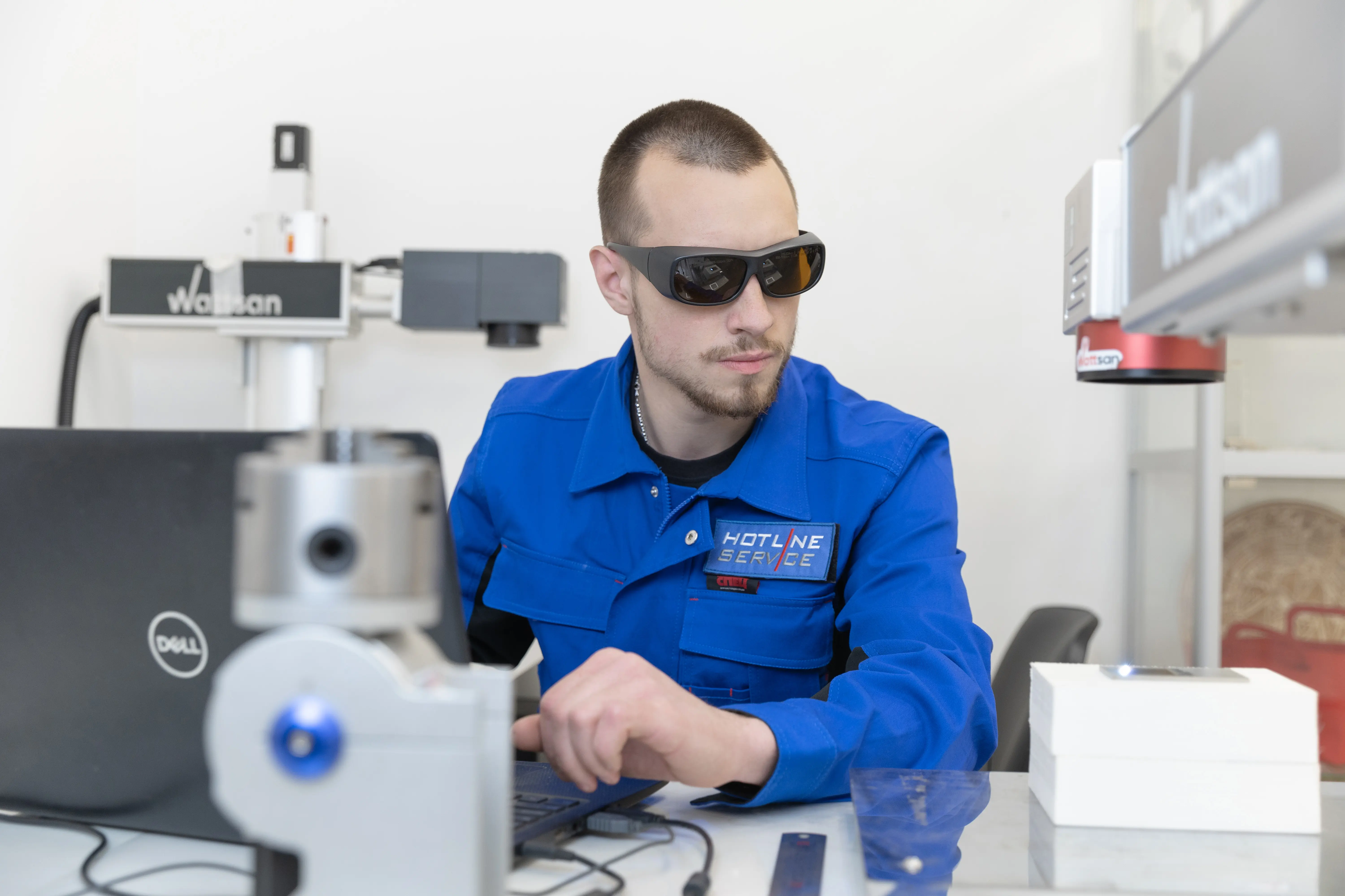 Инженер в защитных лазерных очках и синей рабочей форме управляет лазерным станком и работает за компьютером в лабораторных условиях.